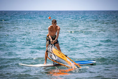 Full length of shirtless man standing on surfboard against sky