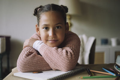 Portrait of smiling girl resting head on arms while sitting at home