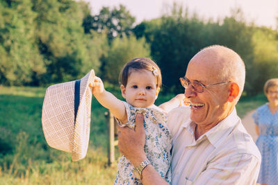 Portrait of granddaughter holding hat with grandfather at park