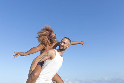 Low angle view of man piggybacking cheerful girlfriend against blue sky