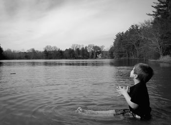 Side view of boy sitting by lake against sky