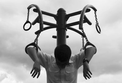 Man hanging from metallic rings against sky