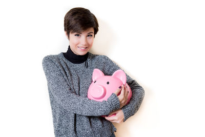 Portrait of smiling young woman holding pink piggy bank against white wall