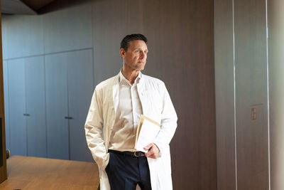 Thoughtful doctor wearing lab coat standing with hand in pocket at hospital