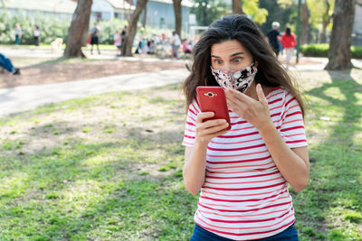 Shocked woman with mask looking at mobile phone while standing in park