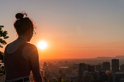 Rear view of woman standing by buildings against sky during sunset