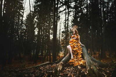 Young woman covered with leaves sitting on tree at forest during autumn