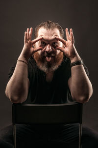 Portrait of man making funny face while sitting on chair against black background