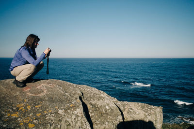 Side view of woman photographing while crouching on rocky shore