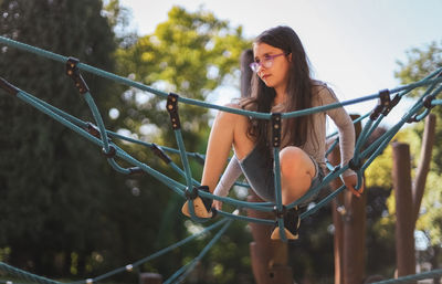 A caucasian girl sits on a rope swing in a park on a playground