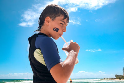 Boy, tattoo of australian flag on cheek, drinking with straw, wearing wetsuit, standing at beach