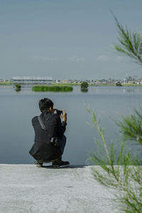 Rear view of boy photographing while crouching by lake against sky