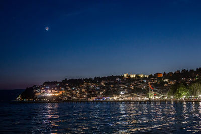 Illuminated city by sea against clear sky at night