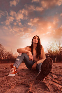 Portrait of young woman sitting on land against sky during sunset