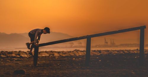 Side view of boy climbing on railing at beach during sunset