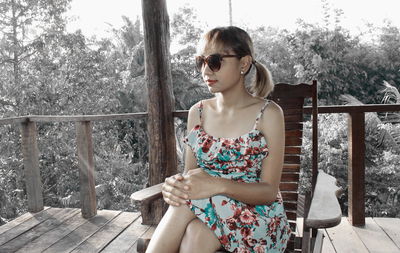 Beautiful young woman looking down while sitting on railing against trees