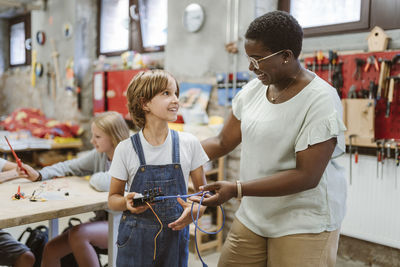 Smiling female teacher encouraging student holding electrical part at technology workshop in school