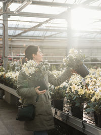 Adult woman in casual clothes buying potted flowers with green leaves and white buds in spacious contemporary greenhouse