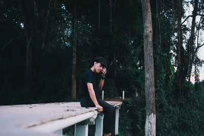Young man sitting on bridge against trees in forest