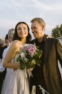 Portrait of happy newlywed woman holding bouquet while standing with father at wedding celebration on rooftop