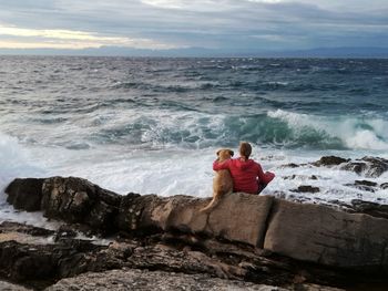 Rear view of woman sitting with dog on rocky shore during sunset