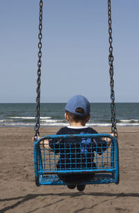 Rear view of boy sitting on swing at beach against clear blue sky