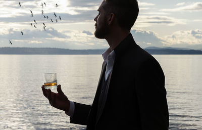 Man holding whiskey glass while standing against sea during sunset