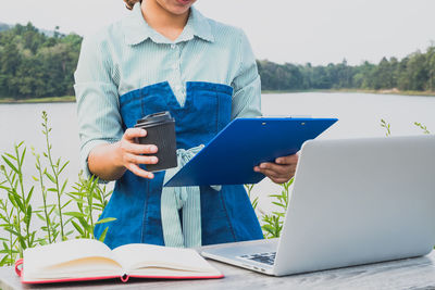 Midsection of woman holding clipboard and coffee cup by laptop and diary on table