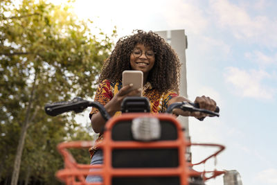 Smiling woman on bicycle using smart phone under sky