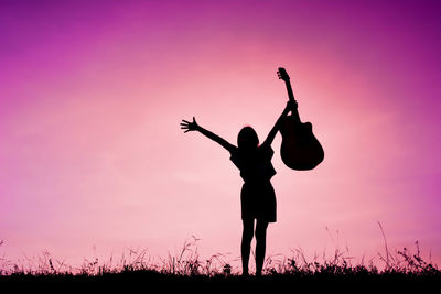Silhouette girl with arms raised holding guitar while standing on field during sunset