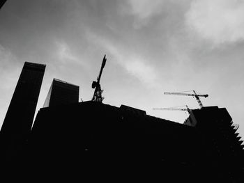 Low angle view of silhouette crane by building against sky