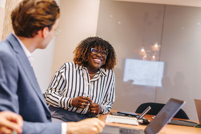 Happy female business professional leaning on table while sitting with colleague in meeting at office