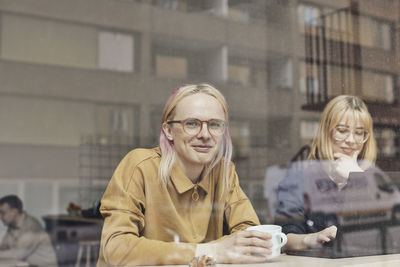 Portrait of smiling computer programmer sitting with female colleague in tech start-up office seen through window