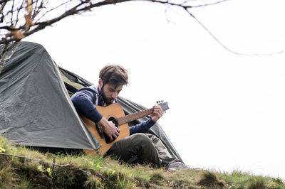 Man playing guitar outside tent on hill against clear sky