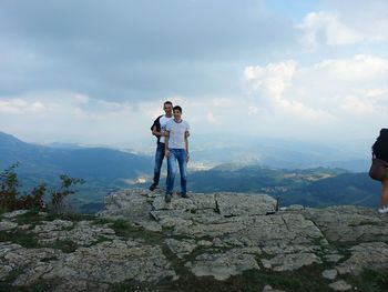 Friends smiling while standing on rock formation against cloudy sky