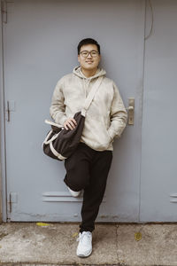 Smiling young man with hand in pocket leaning on gray door