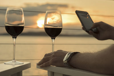 Cropped hands of man with wine glasses using phone by sea against sky during sunset