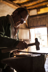 Unrecognizable elderly blacksmith working in a metal sticks with hammer while working in workshop