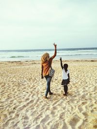 Rear view of woman and girl gesturing peace sign while standing on sand at beach against sky