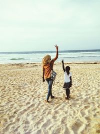 Rear view of woman and girl gesturing peace sign while standing on sand at beach against sky