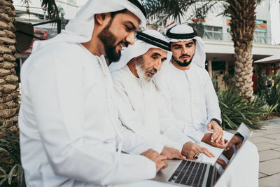 Senior man with grandsons wearing dish dash using laptop outdoors