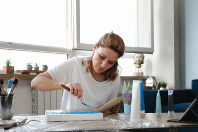 Woman artist painting picture at home with acrylic paints and spatula over the window. adult hobby