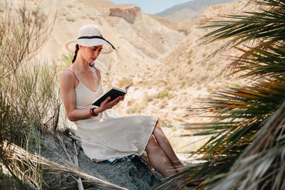 Woman wearing hat reading book while sitting against mountain