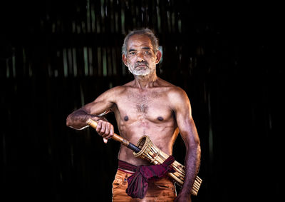 Portrait of shirtless senior man holding weapon standing against black background