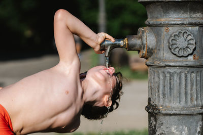 Side view of crop anonymous shirtless male athlete pouring water from tap while working out in park