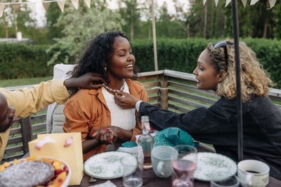 Man and woman looking at necklace of female friend sitting at dining table in back yard