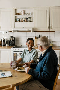 Smiling female nurse talking with senior man having breakfast while sitting at dining table in kitchen