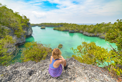 Full length of woman sitting on rock looking at backwater against sky