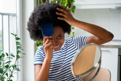 Cool african woman with comb sticking out of curly hair smiling looks in mirror and touches wig