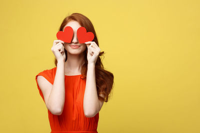 Young woman holding red heart shape paper while standing against yellow background
